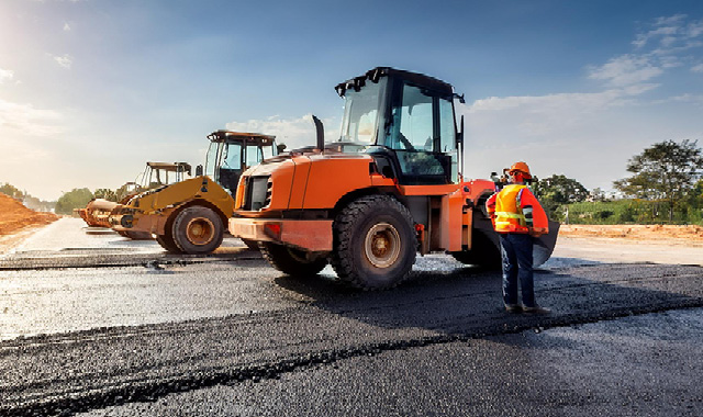 Three construction vehicles line up on a newly paved asphalt road 